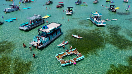 Aerial view of a lively sandbar party with pontoon boats, floating lounge platforms and paddleboards on crystal-clear turquoise water on a sunny summer day.