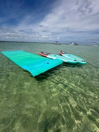 Two striped stand-up paddleboards and a turquoise floating mat anchored in crystal-clear shallow water with boats on the horizon under a bright, partly cloudy sky — beach day scene.