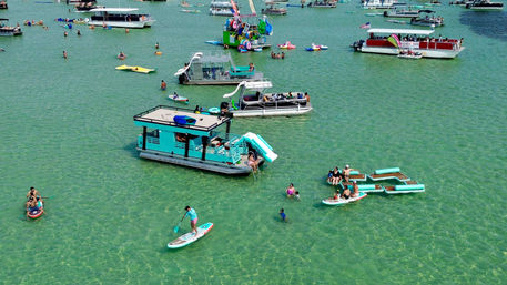 Aerial view of a busy sandbar in clear turquoise water with colorful pontoon boats and houseboats, inflatable slides and rafts, paddleboarders and swimmers enjoying a sunny summer day