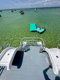 Splash-ready view from a boat deck over clear turquoise shallow water to a small floating dock with two striped paddleboards and scattered boats on a sunny coastal bay