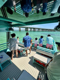 Group of people on a turquoise boat deck gazing at a long concrete bridge over clear green water, with their reflections visible on the glossy roof.
