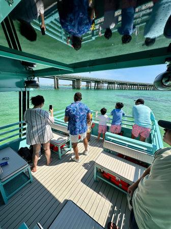 Group of people on a turquoise boat deck gazing at a long concrete bridge over clear green water, with their reflections visible on the glossy roof.