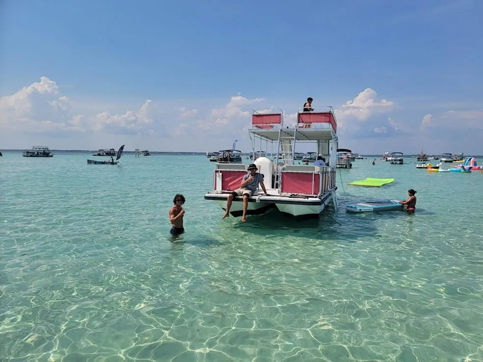 Sunny sandbar scene with a two-level pontoon boat anchored in crystal-clear turquoise shallow water, people swimming and paddling nearby and other boats dotting the horizon.