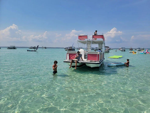 Sunny sandbar scene with a two-level pontoon boat anchored in crystal-clear turquoise shallow water, people swimming and paddling nearby and other boats dotting the horizon.