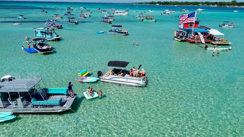 Aerial view of a sunny sandbar party in crystal-clear turquoise water dotted with anchored pontoons, party boats, inflatables and swimmers