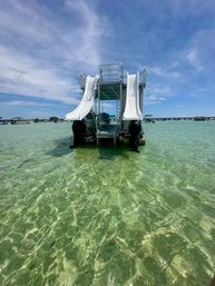 Pontoon boat with twin white water slides anchored in clear shallow turquoise water under a sunny blue sky, with other boats and a distant bridge on the horizon