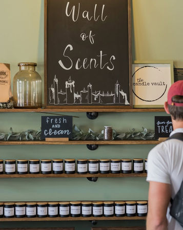 Boutique candle shop interior with a chalkboard reading “Wall of Scents” above wooden shelves lined with labeled scented candle jars, small signs like “fresh and clean,” greenery accents, and a customer in a red cap browsing the display.