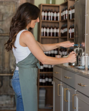 Smiling woman in a green apron fills a glass jar from a stainless-steel spout in a bright artisanal workshop lined with shelves of amber bottles