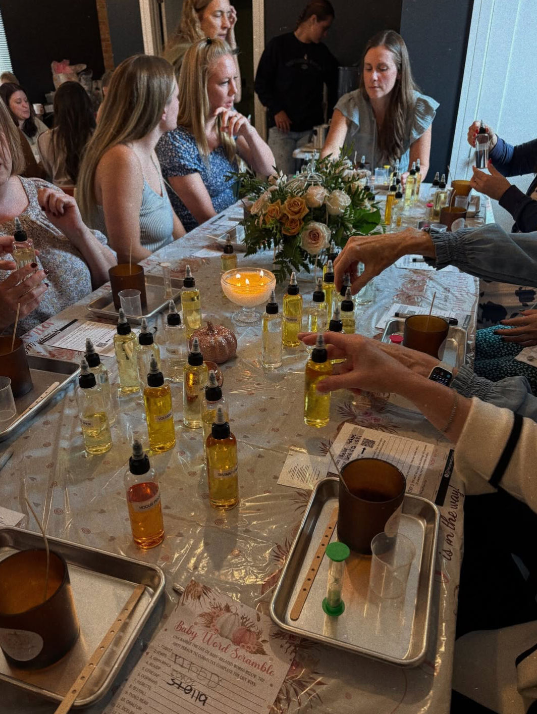 Indoor candle-making party at a long table, participants sampling amber fragrance oils from many dropper bottles, pouring into jars on trays, with a lit candle and rose centerpiece in the middle.