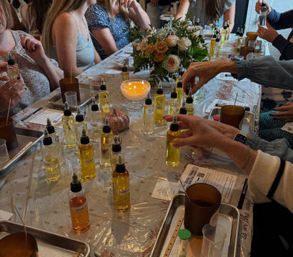 Indoor candle-making party at a long table, participants sampling amber fragrance oils from many dropper bottles, pouring into jars on trays, with a lit candle and rose centerpiece in the middle.