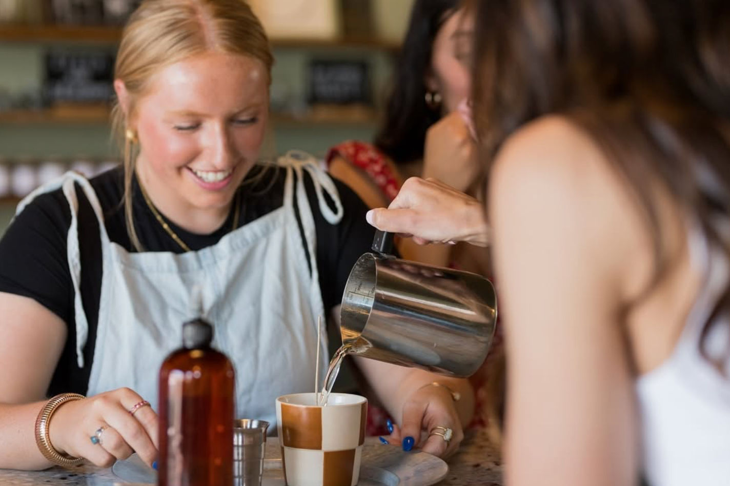 Person pouring steamed milk into a checkered latte cup while smiling friends gather at a cozy coffee shop counter