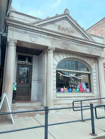 Historic bank façade on a small-town main street, now a candle shop with classical columns and an arched window displaying pastel Easter eggs, garland, and bunny decorations.