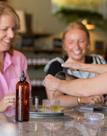 Two smiling women at a DIY candle-making workshop pouring melted wax into a decorative glass jar on a marble countertop, amber refill bottle and tools in the foreground.