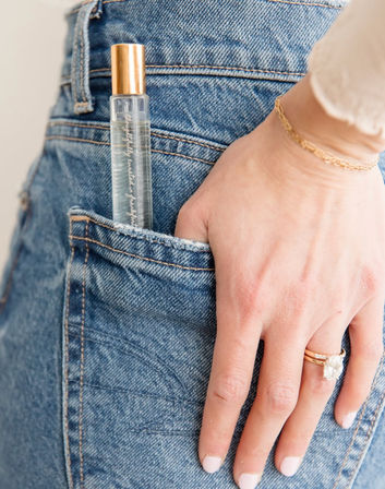 Clear roller perfume bottle with gold cap tucked into blue denim back pocket, close-up of hand wearing gold rings and delicate bracelet