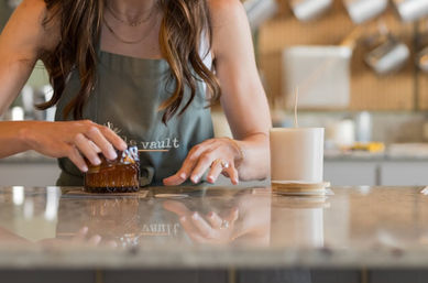 Hands arranging an amber glass candle on a coaster beside a white wax candle on a glossy marble countertop in an artisan candle-making workshop with a blurred kitchen background — handmade candle craft scene.