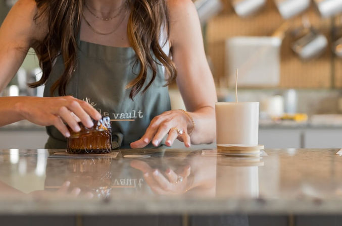 Hands arranging an amber glass candle on a coaster beside a white wax candle on a glossy marble countertop in an artisan candle-making workshop with a blurred kitchen background — handmade candle craft scene.