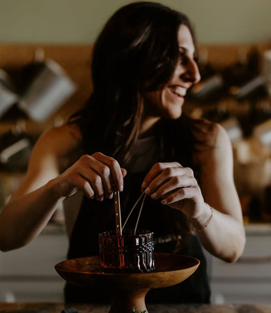 Smiling artisan arranging wicks in a decorative glass jar on a wooden pedestal while making a handmade candle in a cozy kitchen workshop