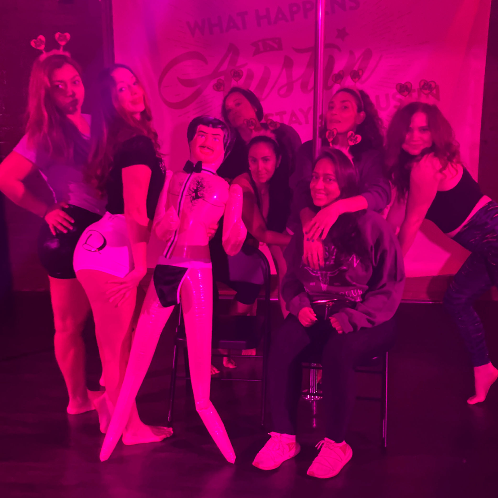Girls' night bachelorette party in a neon-pink dance studio — group of women posing around an inflatable male doll and a pole, some wearing heart headbands.