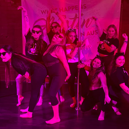 Group of women in a pink-lit dance studio posing playfully around a pole, barefoot and wearing sunglasses.