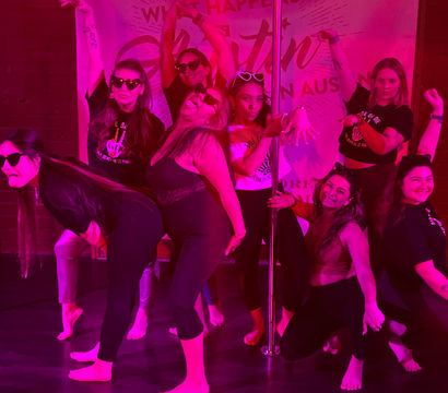 Group of women in a pink-lit dance studio posing playfully around a pole, barefoot and wearing sunglasses.