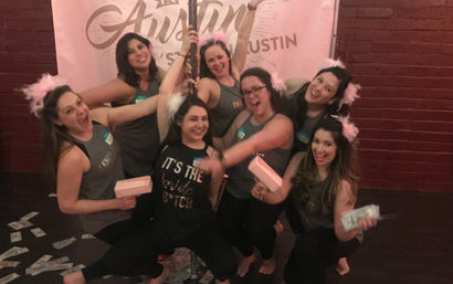 Eight women smiling and cheering around a dance pole at an Austin bachelorette party, wearing matching tanks and pink feather headbands, holding play money and small gift boxes.