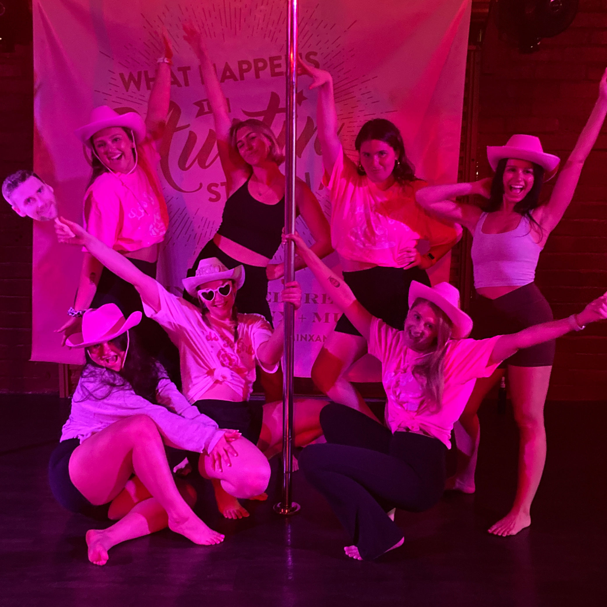 Eight women in cowboy hats and activewear posing around a pole under neon pink lights in a pole-dance studio, smiling and striking celebratory group-fitness poses.