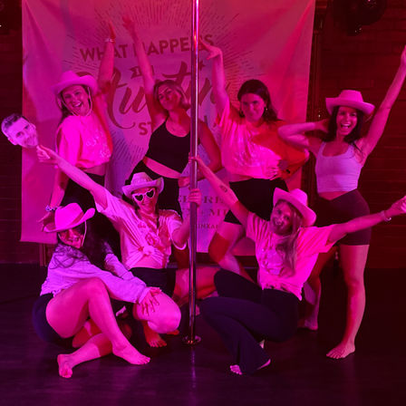 Eight women in cowboy hats and activewear posing around a pole under neon pink lights in a pole-dance studio, smiling and striking celebratory group-fitness poses.