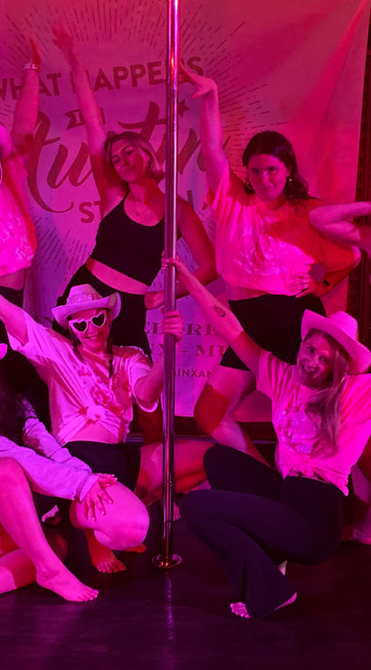 Eight women in cowboy hats and activewear posing around a pole under neon pink lights in a pole-dance studio, smiling and striking celebratory group-fitness poses.
