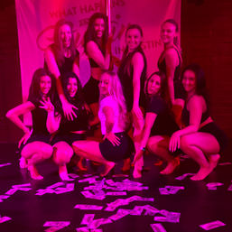 Nine women in dancewear posing around a pole on a nightclub stage under pink lighting, with dollar bills scattered across the floor for a festive party vibe.