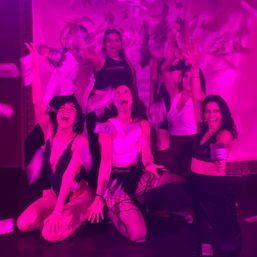 Group of women on a nightclub stage under pink lighting, cheering and tossing dollar bills during a lively dance/party performance.