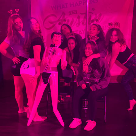 Group of women in neon pink club lighting posing around an inflatable tuxedo doll and pole, wearing heart headbands for a bachelorette-style girls' night