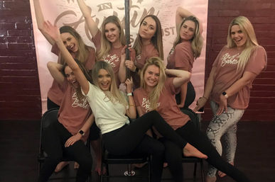 Eight women in matching rose t-shirts and leggings posing around a pole in a brick-walled dance studio, smiling during a bachelorette celebration.