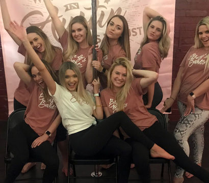 Eight women in matching rose t-shirts and leggings posing around a pole in a brick-walled dance studio, smiling during a bachelorette celebration.