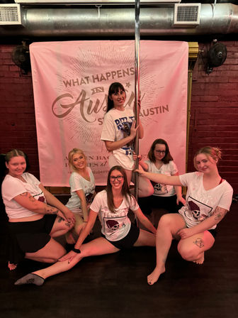 Smiling group of women in matching bachelorette shirts posing around a chrome pole in front of a pink "What Happens in Austin" backdrop during an Austin bachelorette pole-dance party