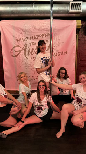 Smiling group of women in matching bachelorette shirts posing around a chrome pole in front of a pink "What Happens in Austin" backdrop during an Austin bachelorette pole-dance party