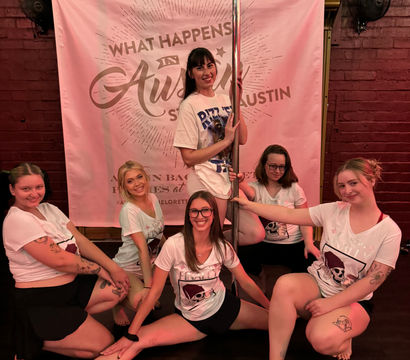 Smiling group of women in matching bachelorette shirts posing around a chrome pole in front of a pink "What Happens in Austin" backdrop during an Austin bachelorette pole-dance party