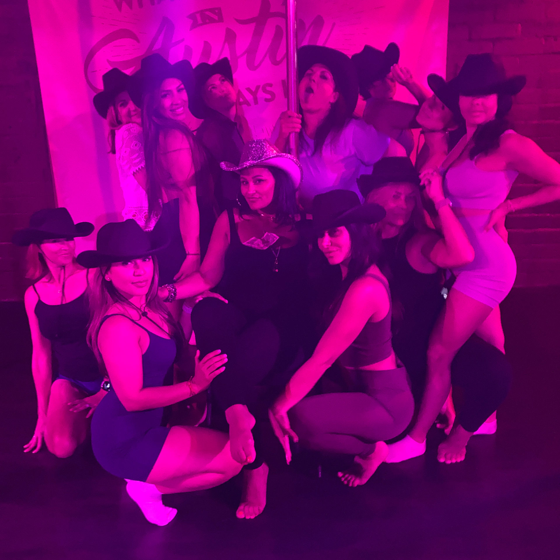 Group of women wearing cowgirl hats posing around a dance pole under pink neon lighting, playful bachelorette or girls' night out vibe in a nightclub/dance studio