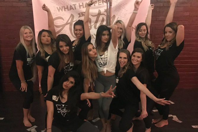 Cheerful bachelorette party group of women striking playful poses around a pole in a pink-decorated Austin dance studio, wearing casual workout clothes with scattered play money on the floor.