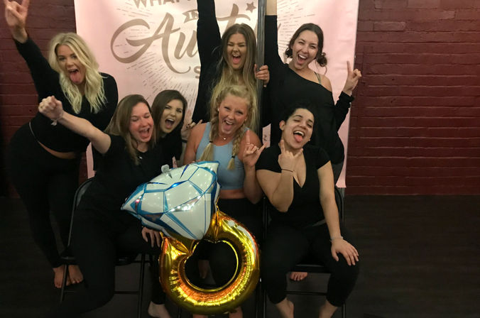 Cheerful group of women posing and celebrating indoors, holding a giant gold ring and diamond-shaped balloon in front of a pink banner and red brick wall.