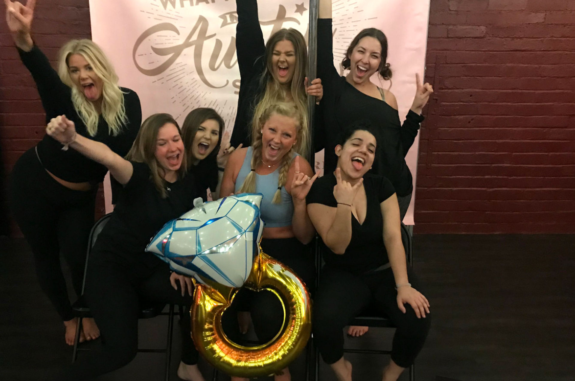 Cheerful group of women posing and celebrating indoors, holding a giant gold ring and diamond-shaped balloon in front of a pink banner and red brick wall.