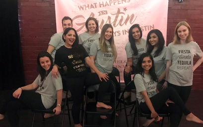 Group of friends posing indoors in front of a pink “What happens in Austin” banner, wearing matching “Fiesta Siesta Tequila Repeat” shirts and black leggings while seated on folding chairs, smiling and having fun.