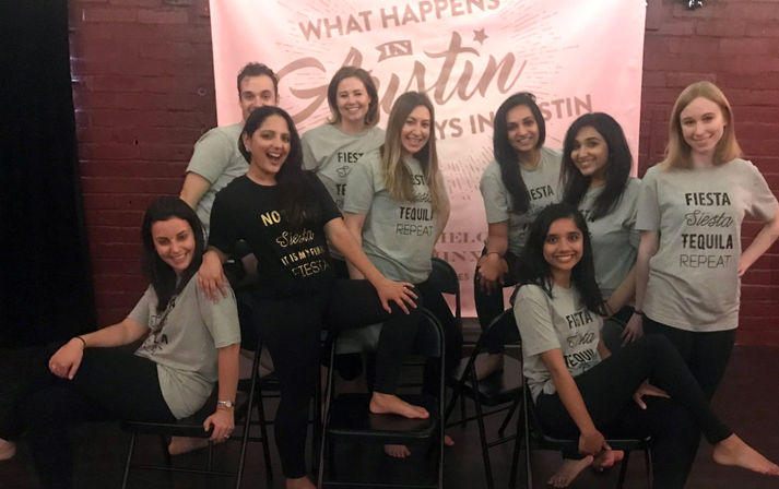 Group of friends posing indoors in front of a pink “What happens in Austin” banner, wearing matching “Fiesta Siesta Tequila Repeat” shirts and black leggings while seated on folding chairs, smiling and having fun.