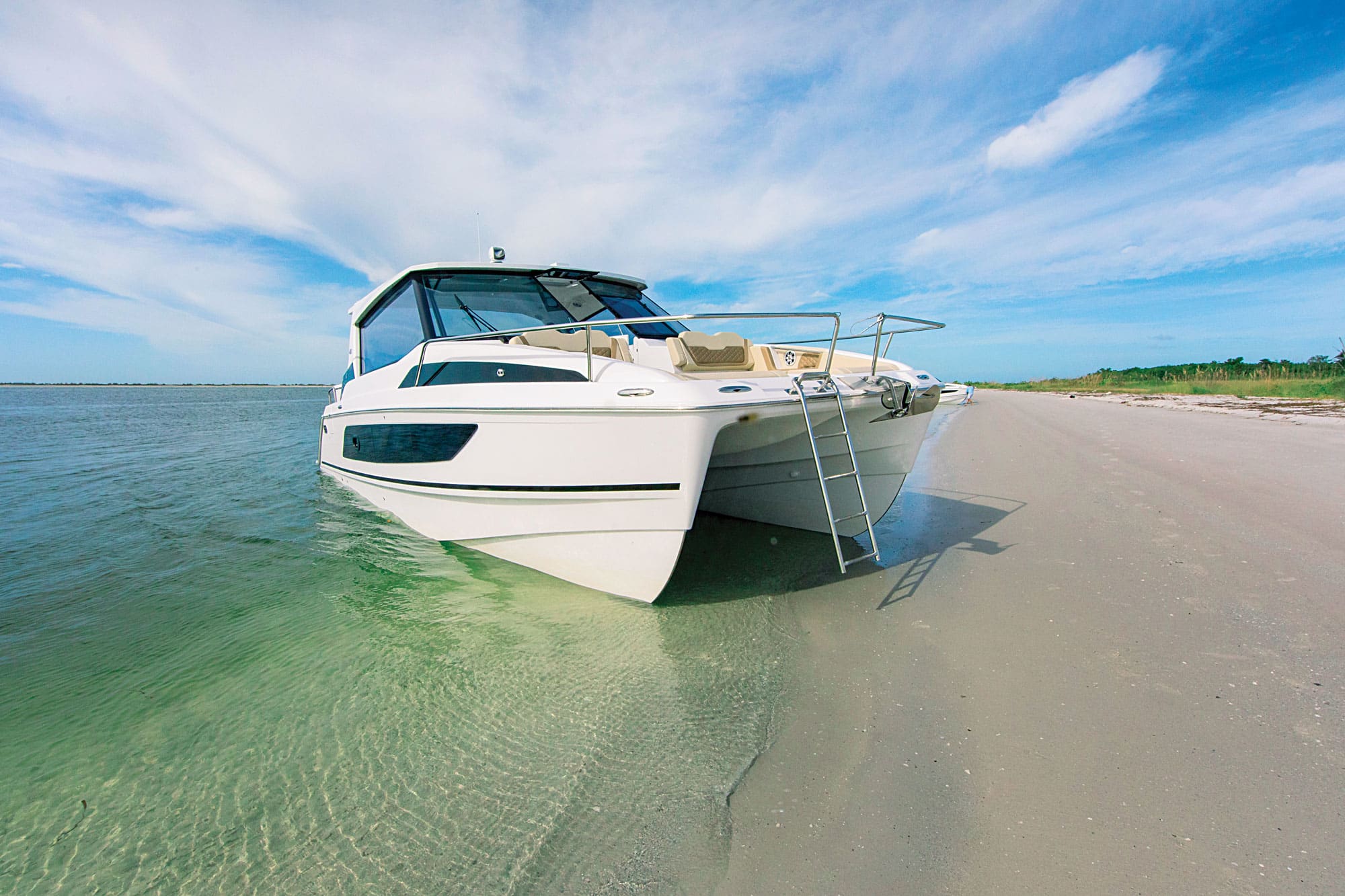 White motor yacht beached in clear shallow turquoise water beside a wide sandy shore, swim ladder down and bright blue sky with wispy clouds.