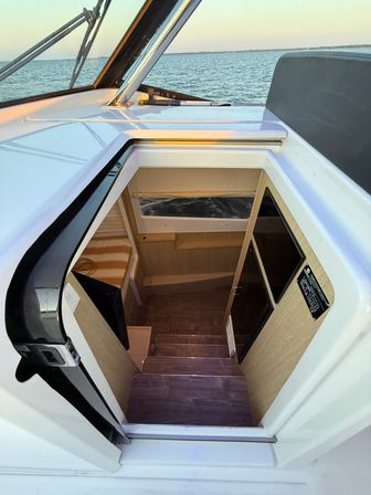Open hatch on a motorboat leading down wooden steps into a cozy cabin with a striped bench, cabin window, and calm ocean at sunset.