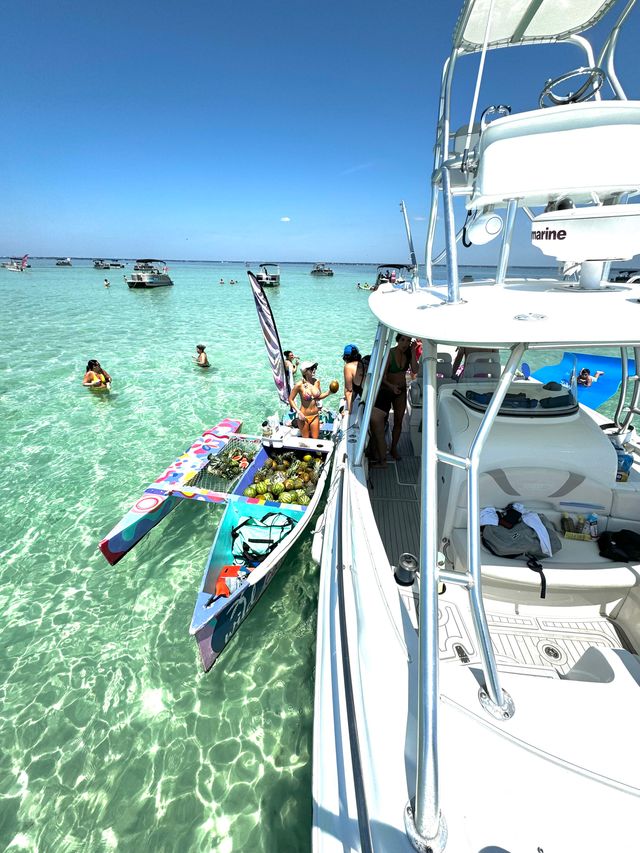 White powerboat anchored at a shallow turquoise sandbar, colorful canoe vendor piled with pineapples, people wading in clear water and boats dotting the horizon under a bright blue sky.