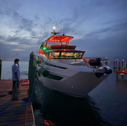 Sleek motor yacht moored at a marina at dusk, red LED canopy lights glowing, people on deck and dock, calm water and dramatic twilight sky.