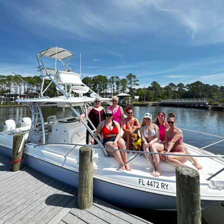 Group of women in colorful swimsuits and a small dog posing on the bow of a white center-console boat at a sunny Florida marina with pine-lined shoreline