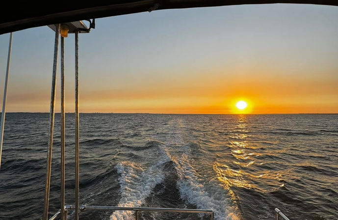 View from a boat stern at sunset: golden sun on the horizon reflecting on rippling open water, foamy wake trailing behind the boat with metal railings and canopy framing the scene.