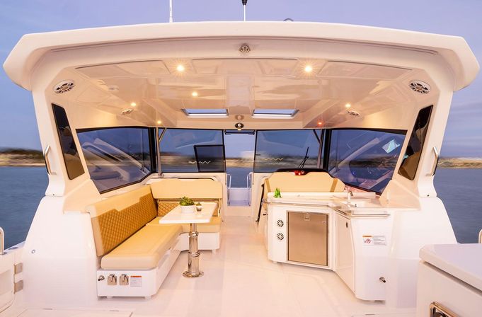 Inviting luxury yacht cockpit at dusk with cushioned L-shaped seating, dining table, compact galley and panoramic windows overlooking calm coastal waters.