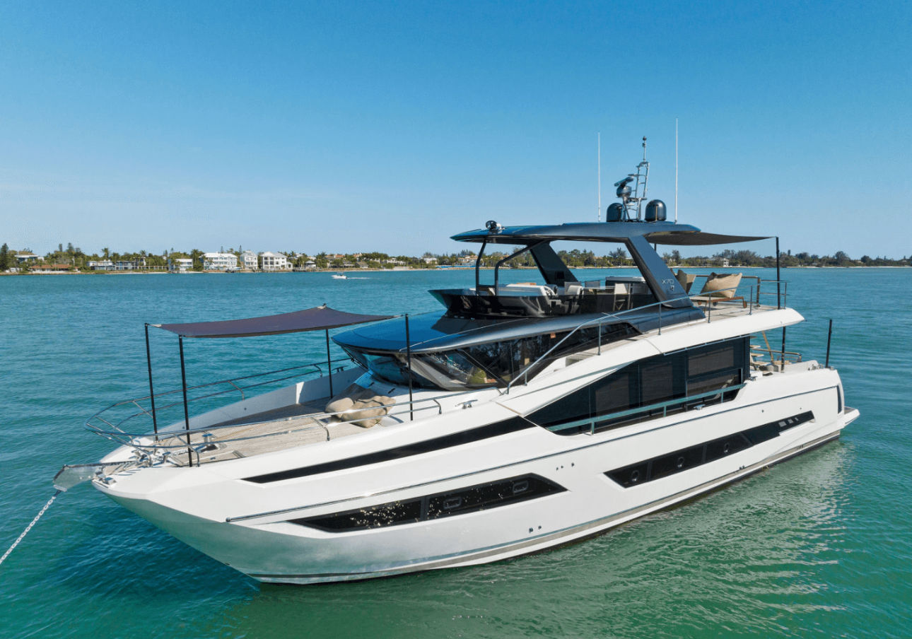 Sleek white luxury motor yacht anchored off a coastal shoreline on turquoise water under a clear blue sky, multi-level sun deck with shaded canopy and seaside homes in the distance.